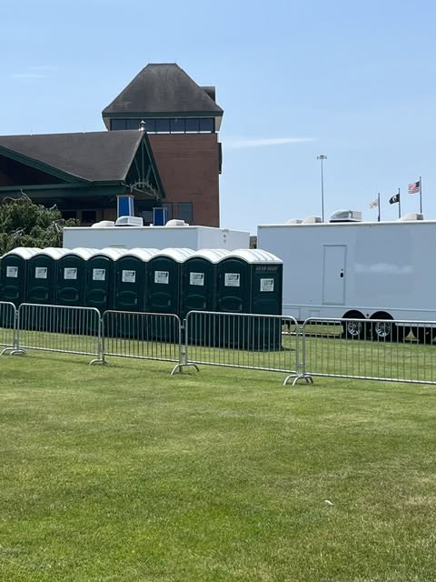 Bank of green portable toilets lined up beside white multi-stall restroom trailers behind crowd control barricades on a field