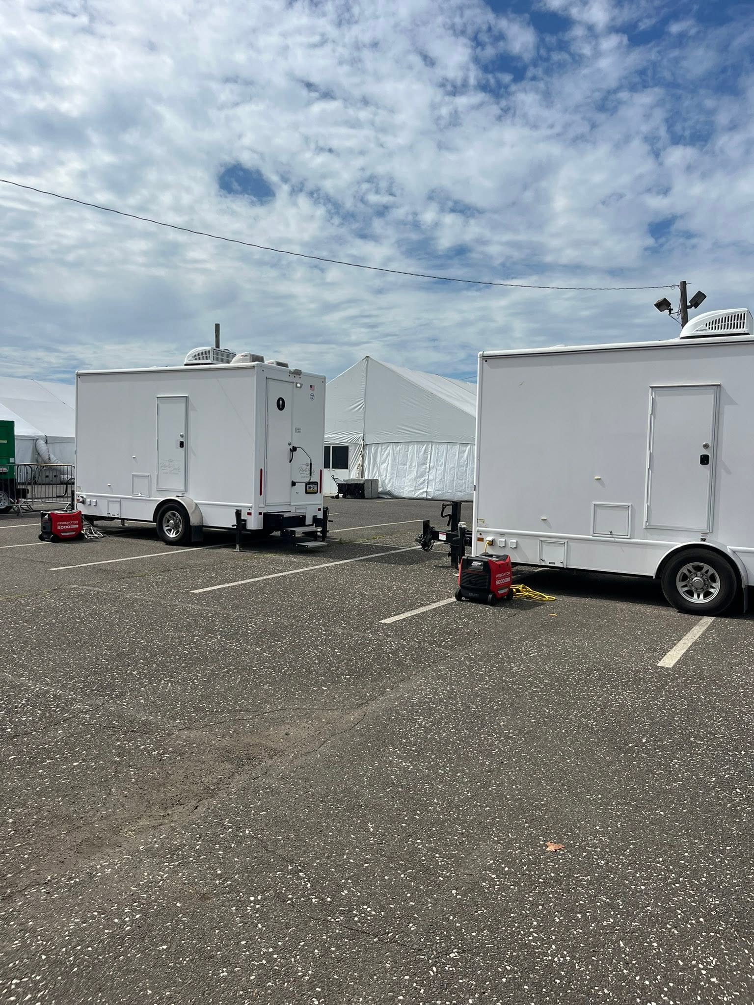 Two white restroom trailers staged on asphalt lot with red power generators next to an outdoor event tent