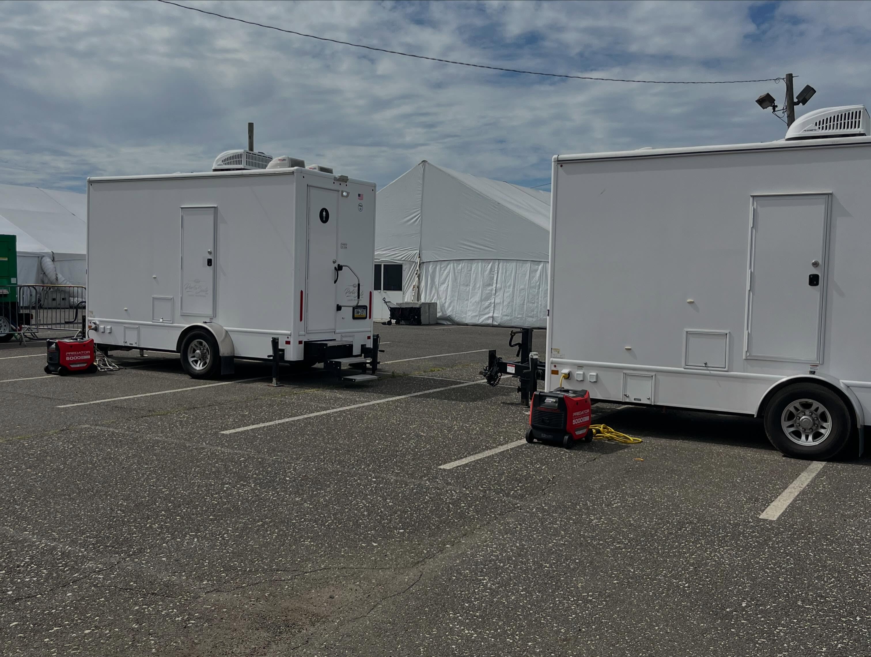 Two white restroom trailers staged on an asphalt lot beside a large white event tent with power generators