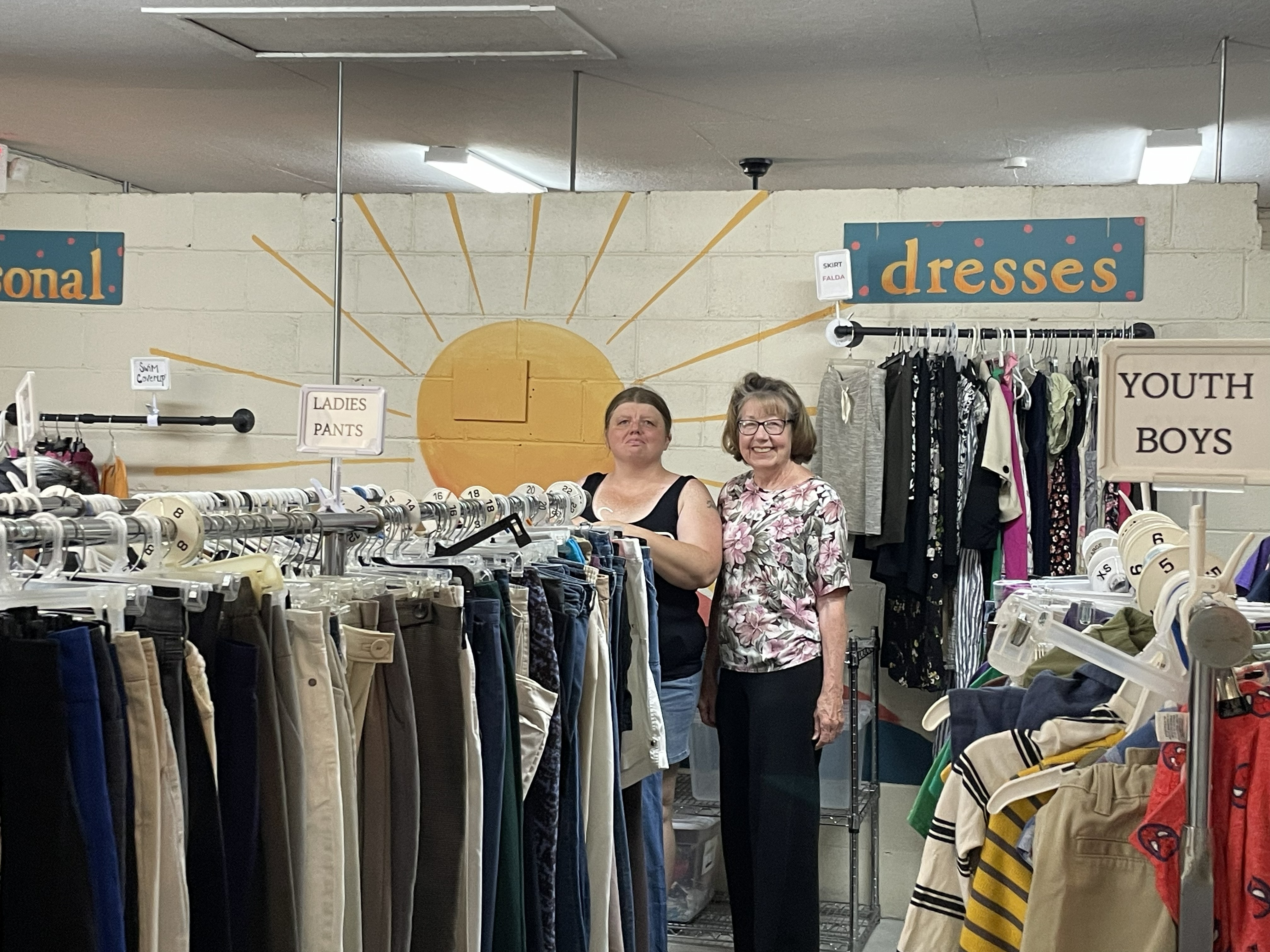 Volunteers in the We Care We Share Ministries clothing closet with organized racks for dresses, pants, youth, and boys sections