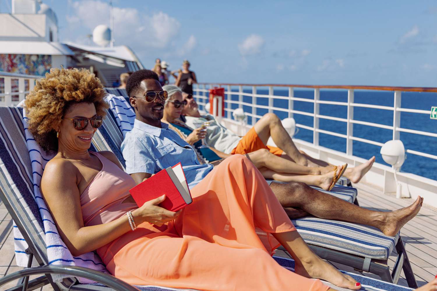 Adult travelers relaxing in cushioned deck loungers reading and enjoying drinks on a sunny cruise deck