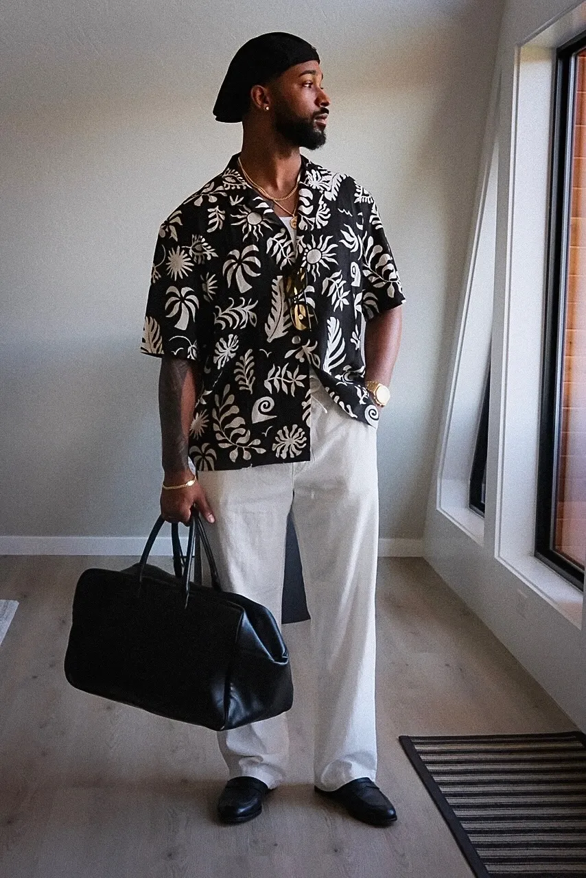 Jai'koa Parham in a black and white patterned resort shirt and white linen trousers holding a leather duffel bag, lifestyle campaign