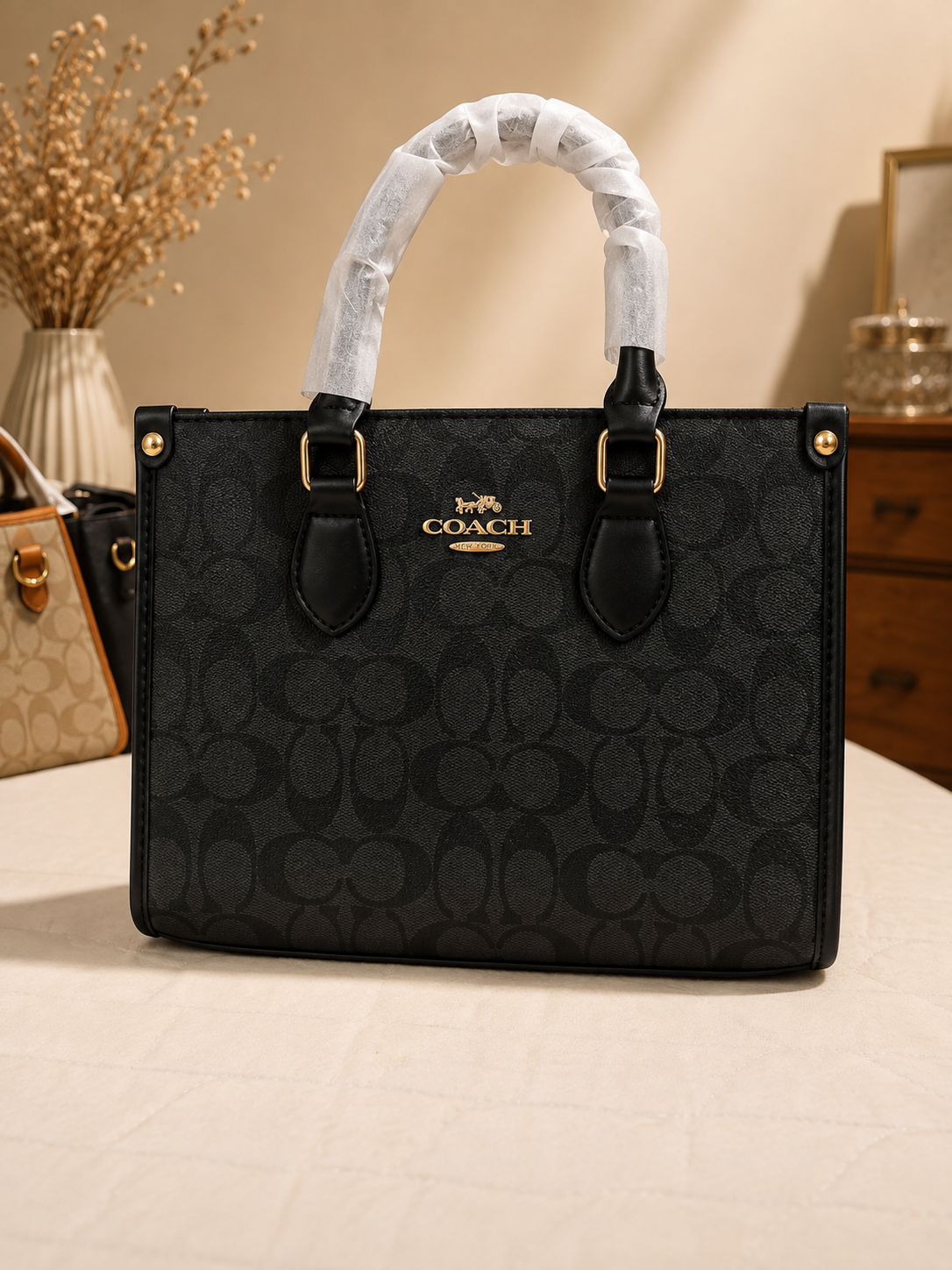 Black Coach signature monogram tote bag with dual top handles gold hardware and protective handle wrapping displayed with other bags in background