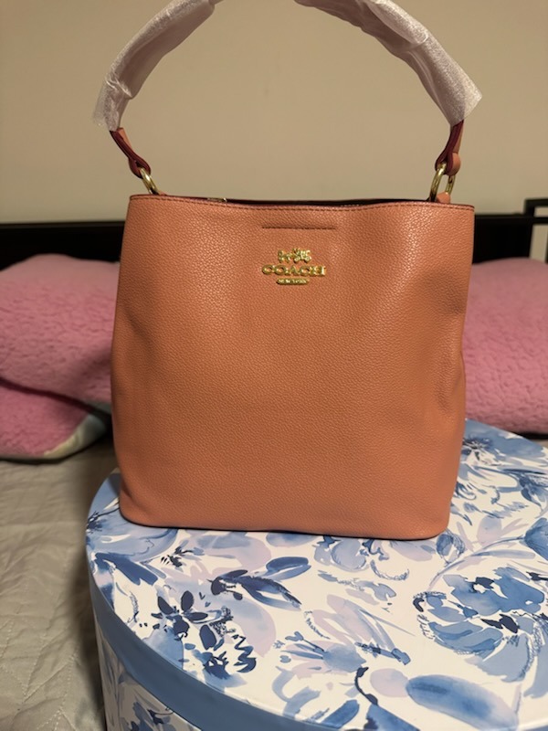 Peach coral Coach leather bucket bag with gold horse carriage logo dual wrapped top handles gold hardware displayed on blue white floral patterned round box with pink blanket background