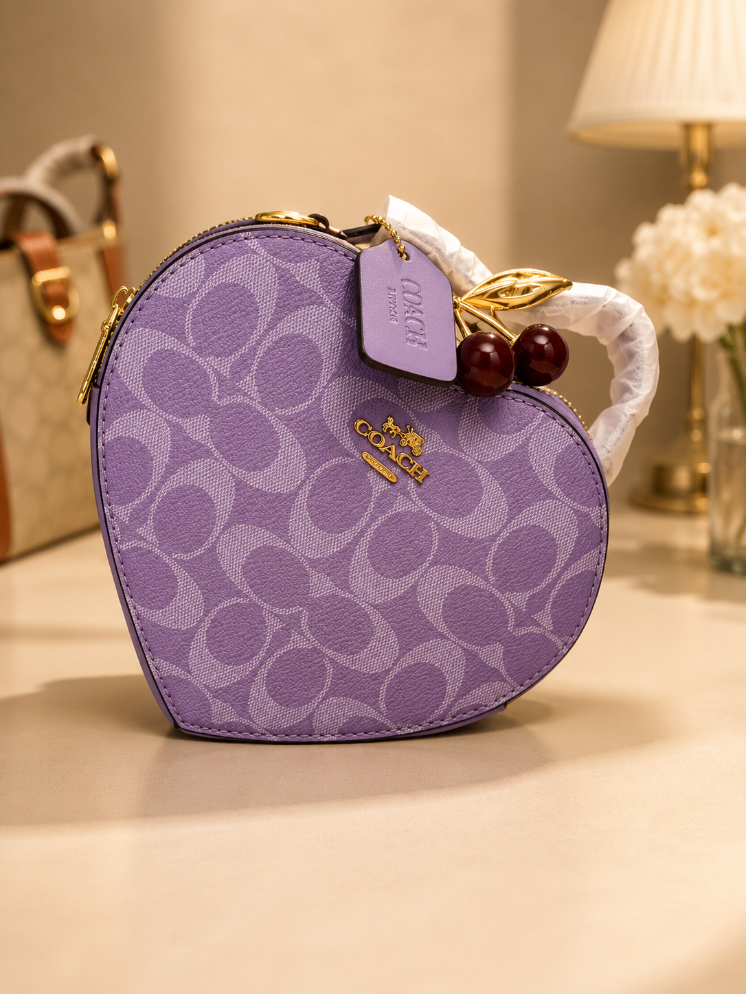 Coach purple heart-shaped signature monogram bag with gold cherry charm keychain white wrapped handle zipper closure and COACH tag on neutral surface with other luxury bags visible