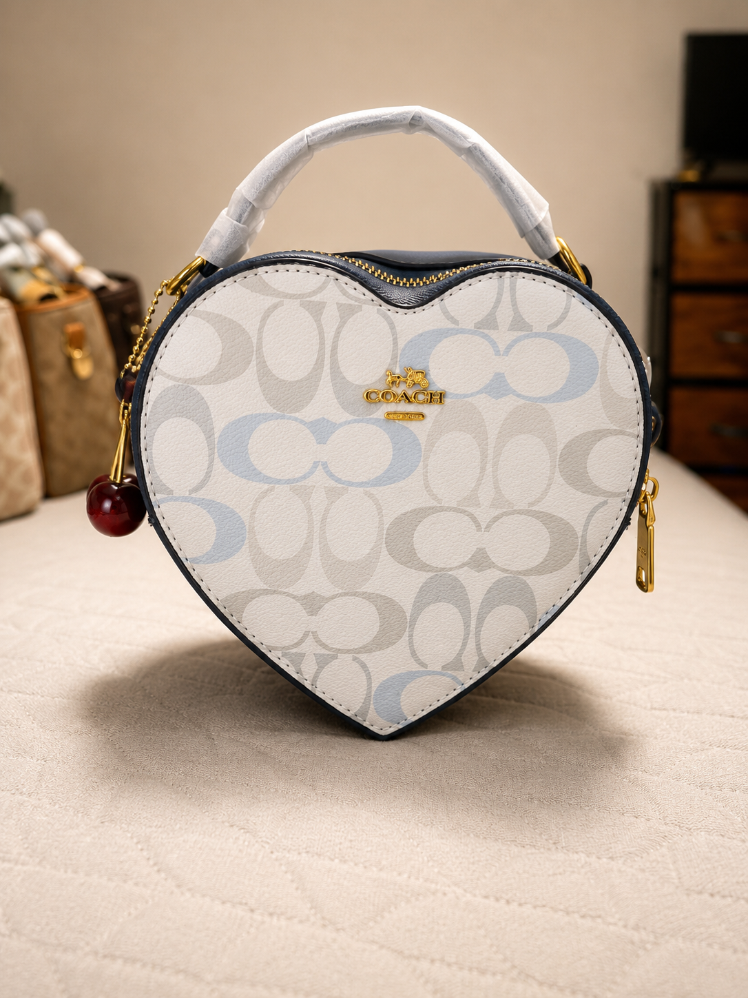 Coach white cream signature monogram heart-shaped bag with light blue accents gold cherry charm keychain white wrapped handle zipper closure and other luxury bags in background