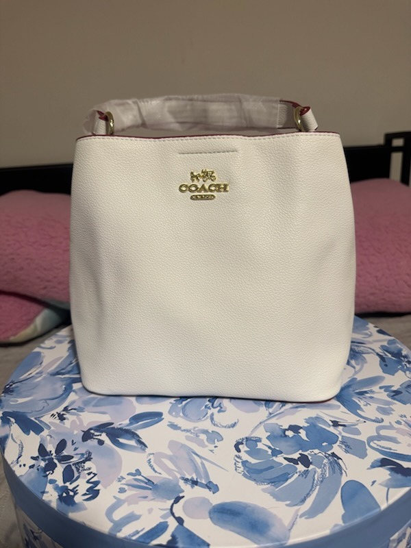 White cream Coach leather handbag with gold COACH logo embossed dual top handles protective wrapping structured minimalist design displayed on blue floral surface