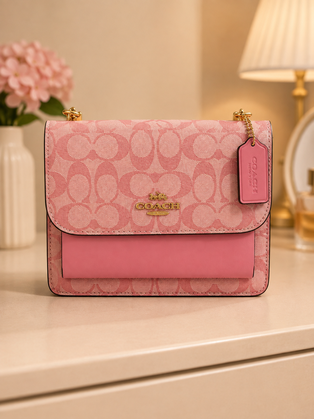 Coach pink signature monogram shoulder bag with gold hardware chain strap and COACH tag displayed on elegant surface with pink flowers and lamp in background