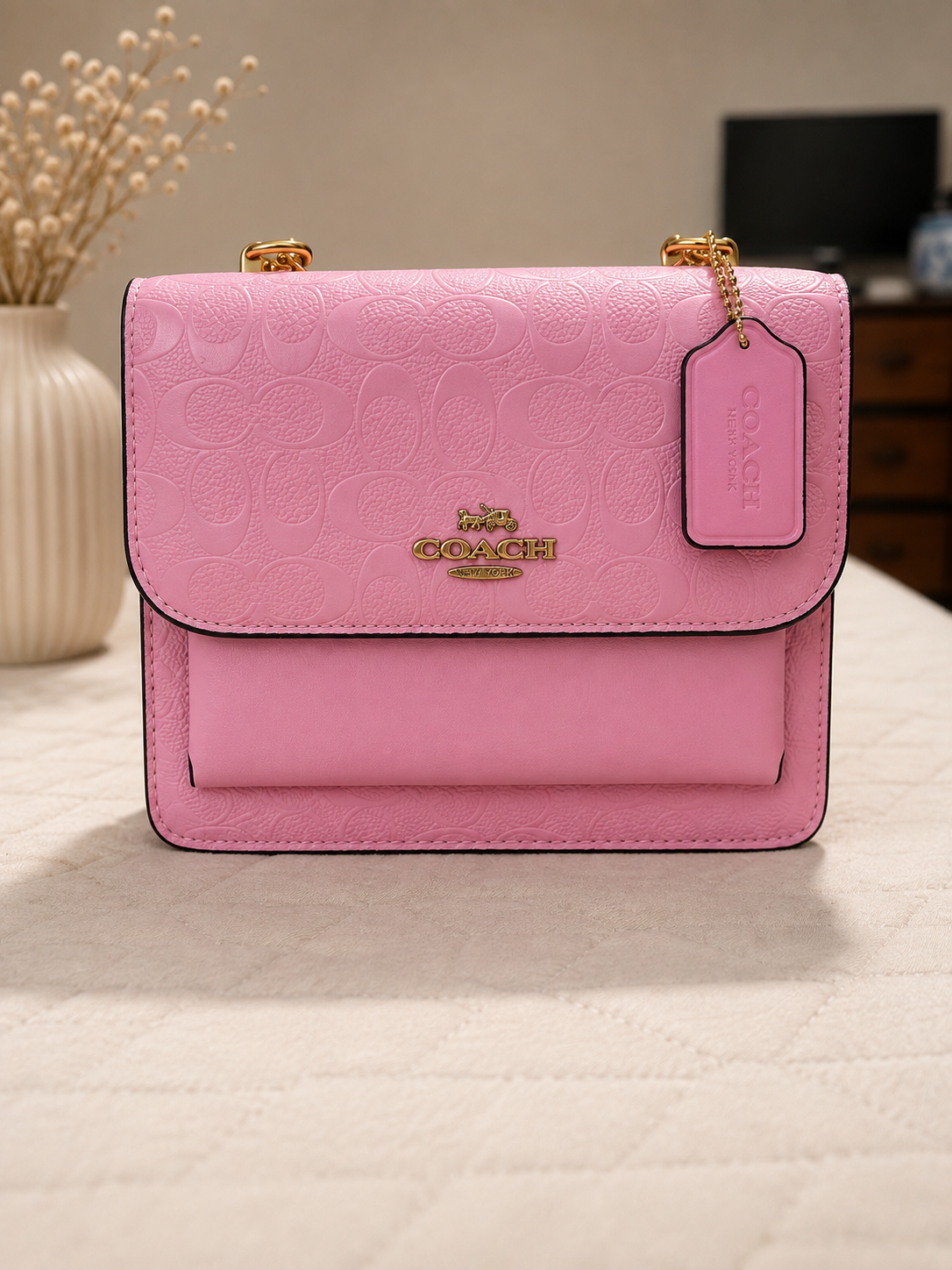 Coach pink embossed leather signature crossbody bag with gold horse and carriage logo front pocket chain strap and leather tag on neutral surface with decorative dried flowers background