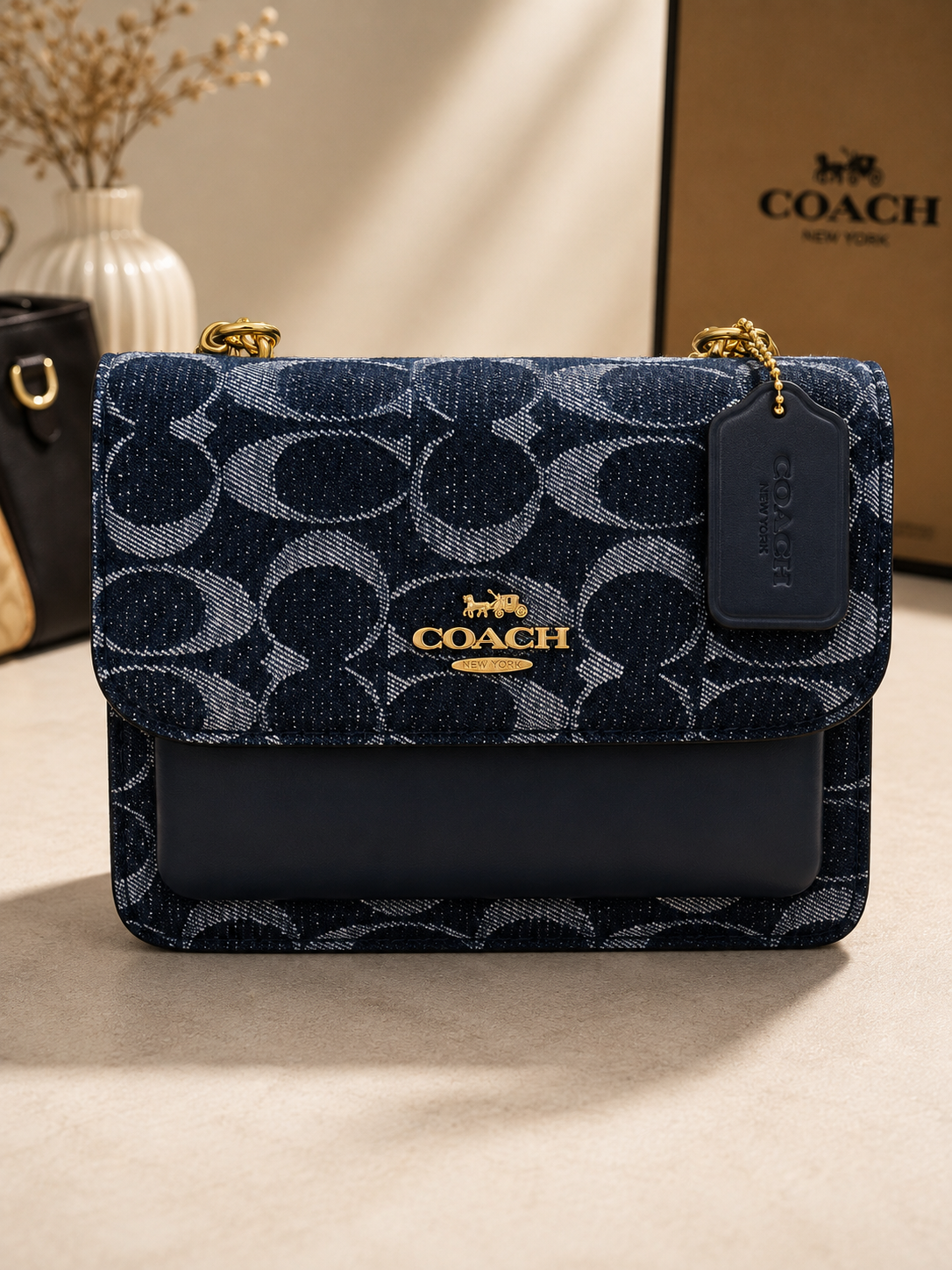 Coach navy blue denim signature monogram shoulder bag with gold hardware chain strap leather tag and structured flap closure displayed on neutral surface with COACH box in background