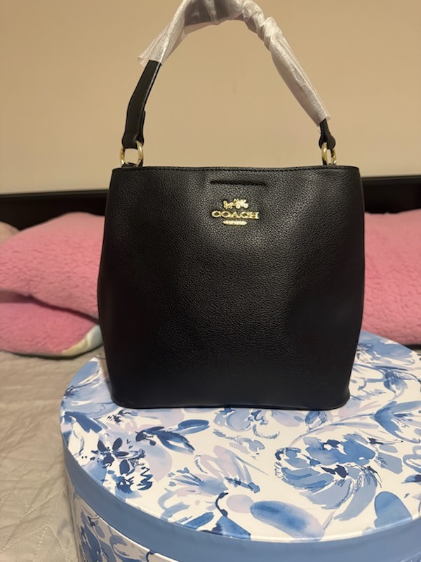 Black Coach leather handbag with gold COACH logo embossed dual top handles protective wrapping structured elegant design displayed on blue floral patterned box