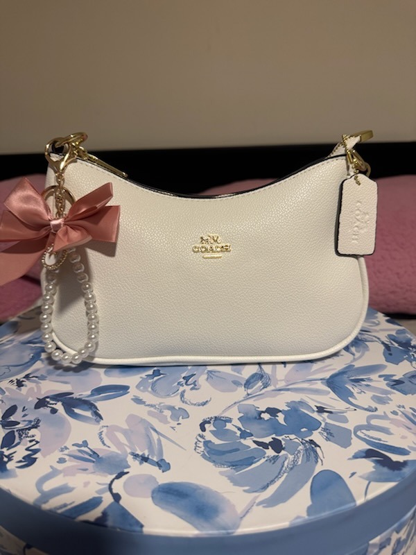 White cream Coach leather shoulder bag with pink satin bow charm pearl beaded chain gold hardware COACH logo tag displayed on elegant blue floral box surface