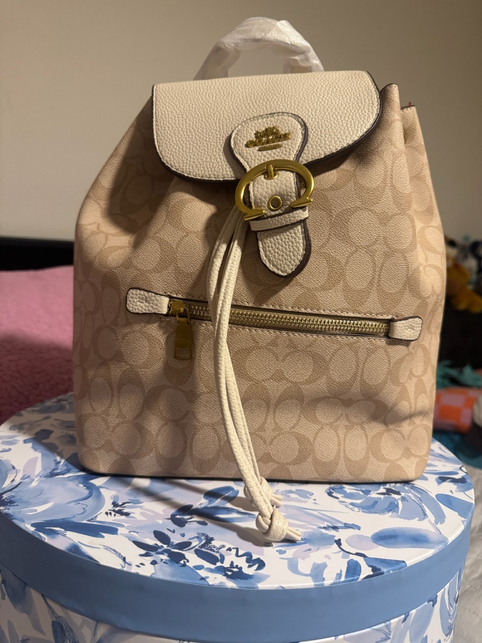 Cream beige Coach signature monogram backpack with white leather top panel gold horse carriage logo buckle drawstring closure front zip pocket on floral display box with pink background