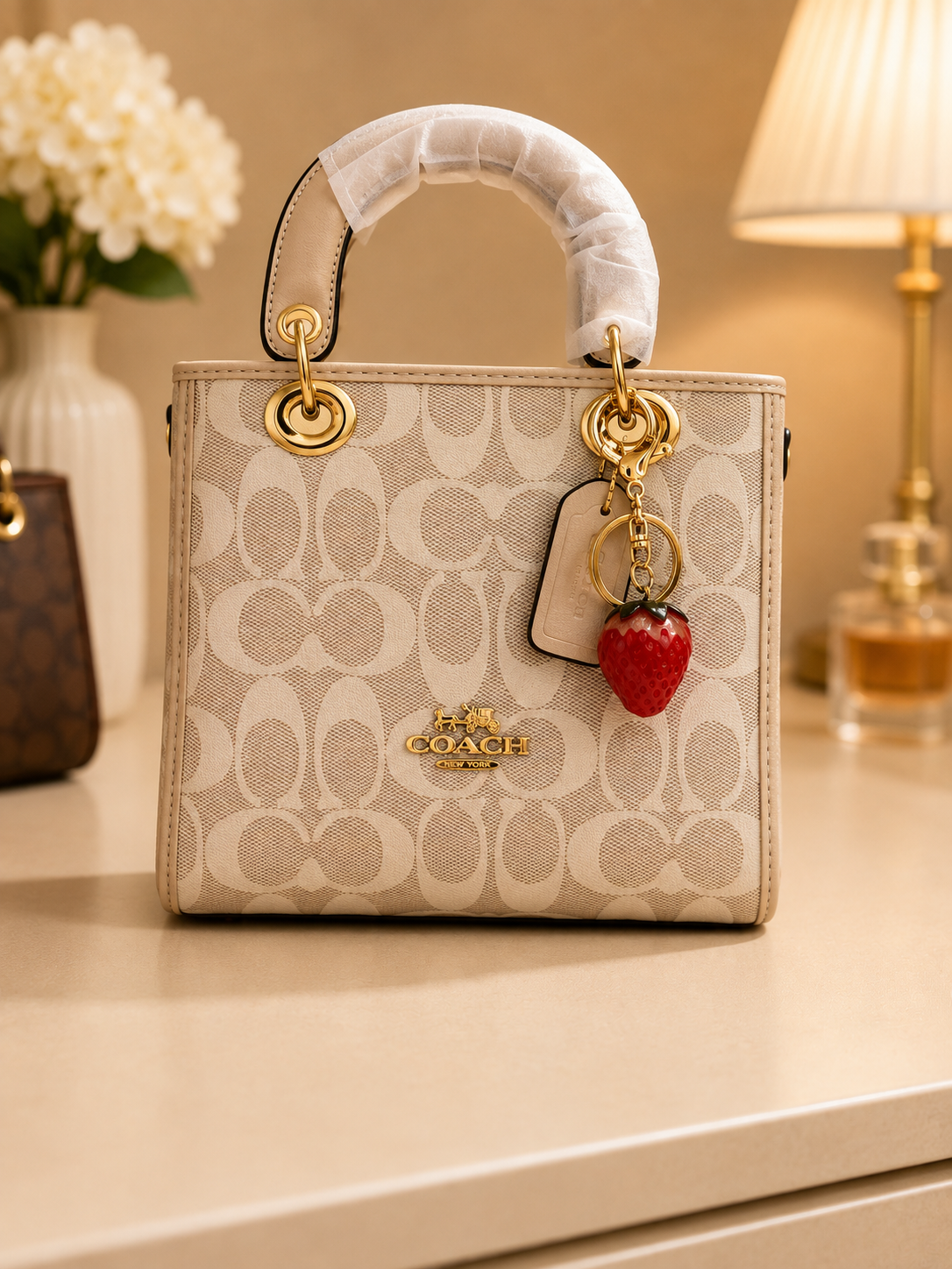 Coach beige signature monogram tote bag with gold ring hardware strawberry red fruit charm keychain leather tag and curved top handle displayed on surface with white flowers perfume bottle in background