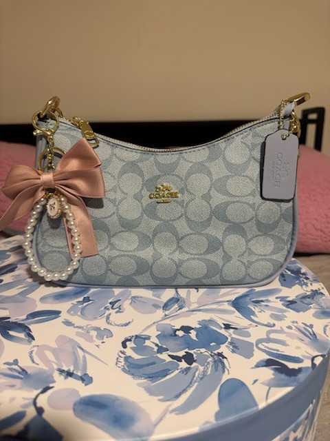 Light blue Coach signature monogram shoulder bag with pink satin bow charm pearl beaded chain gold zipper hardware COACH tag displayed on elegant blue white floral patterned round box