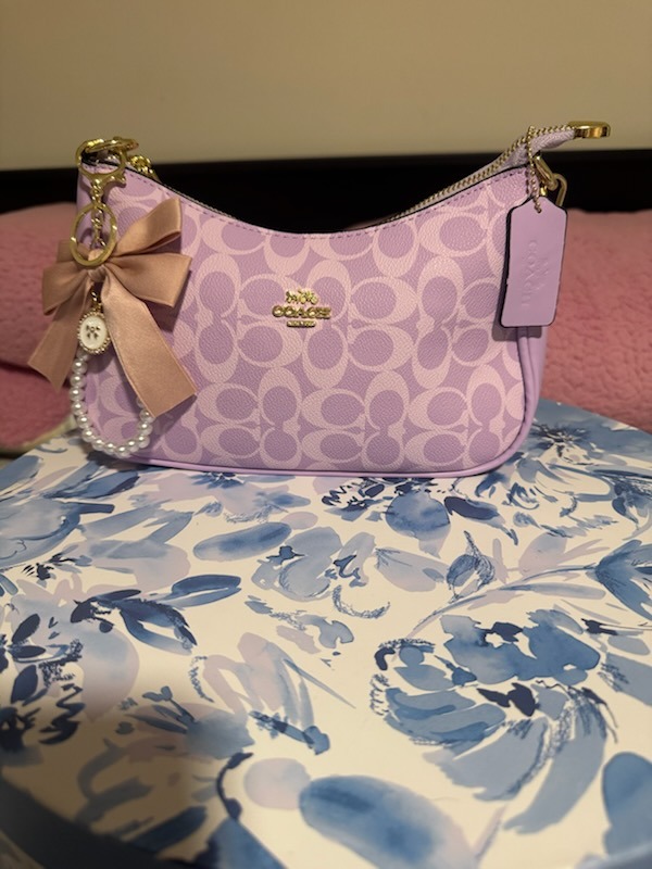 Lavender purple Coach signature monogram shoulder bag with pink satin bow charm pearl beaded chain gold zipper hardware COACH tag on floral display box surface