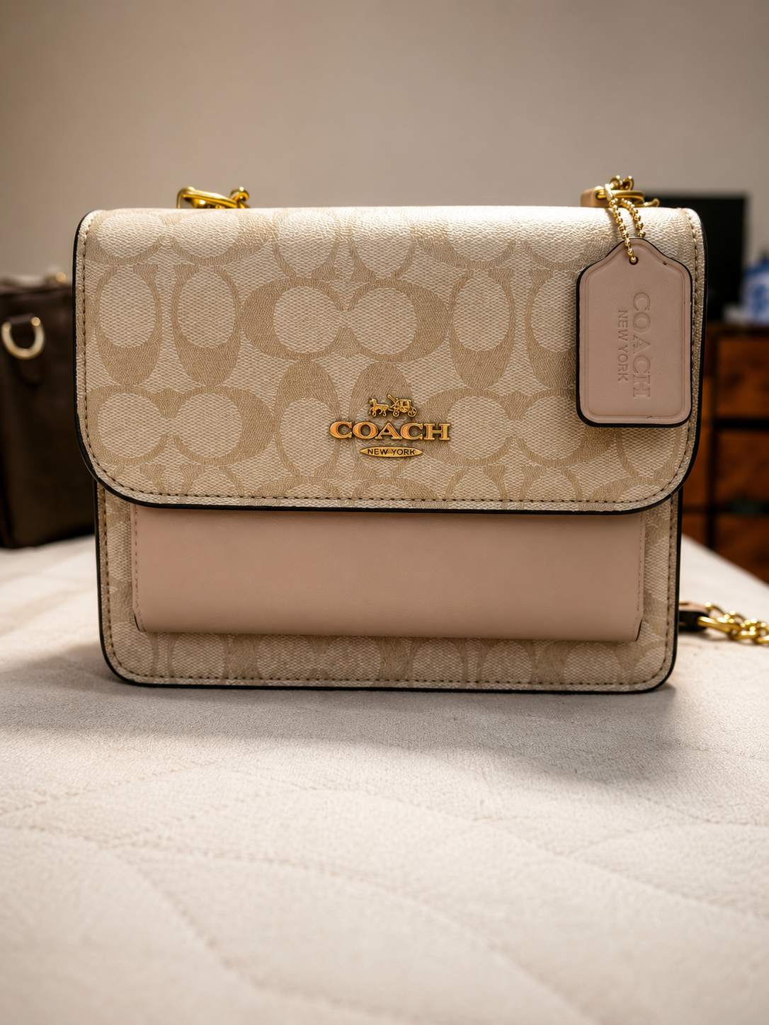 Coach beige signature monogram crossbody bag with gold horse and carriage logo embossed leather front pocket chain strap and leather tag displayed on neutral surface