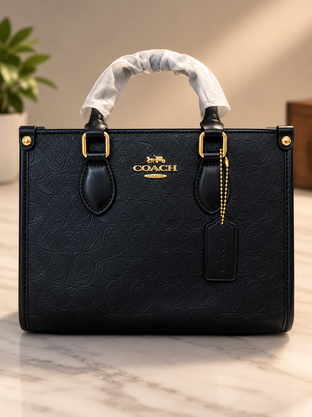 Coach black embossed leather signature handbag with gold hardware dual top handles leather tag with beaded chain strap and protective handle wrapping on marble surface with plant background