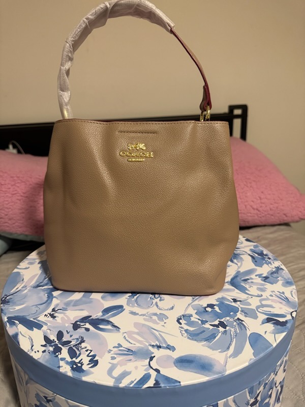 Beige tan Coach leather handbag with gold horse carriage logo embossed dual top handles protective wrapping structured tote design displayed on floral box