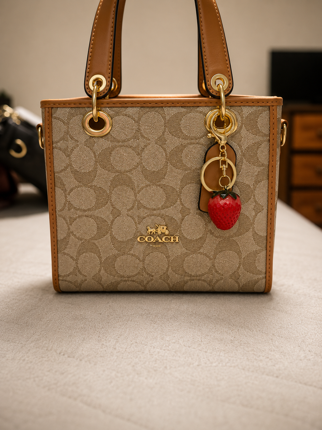 Coach beige tan signature monogram handbag with brown leather trim dual top handles gold ring hardware red strawberry fruit charm keychain and leather tag displayed on neutral surface