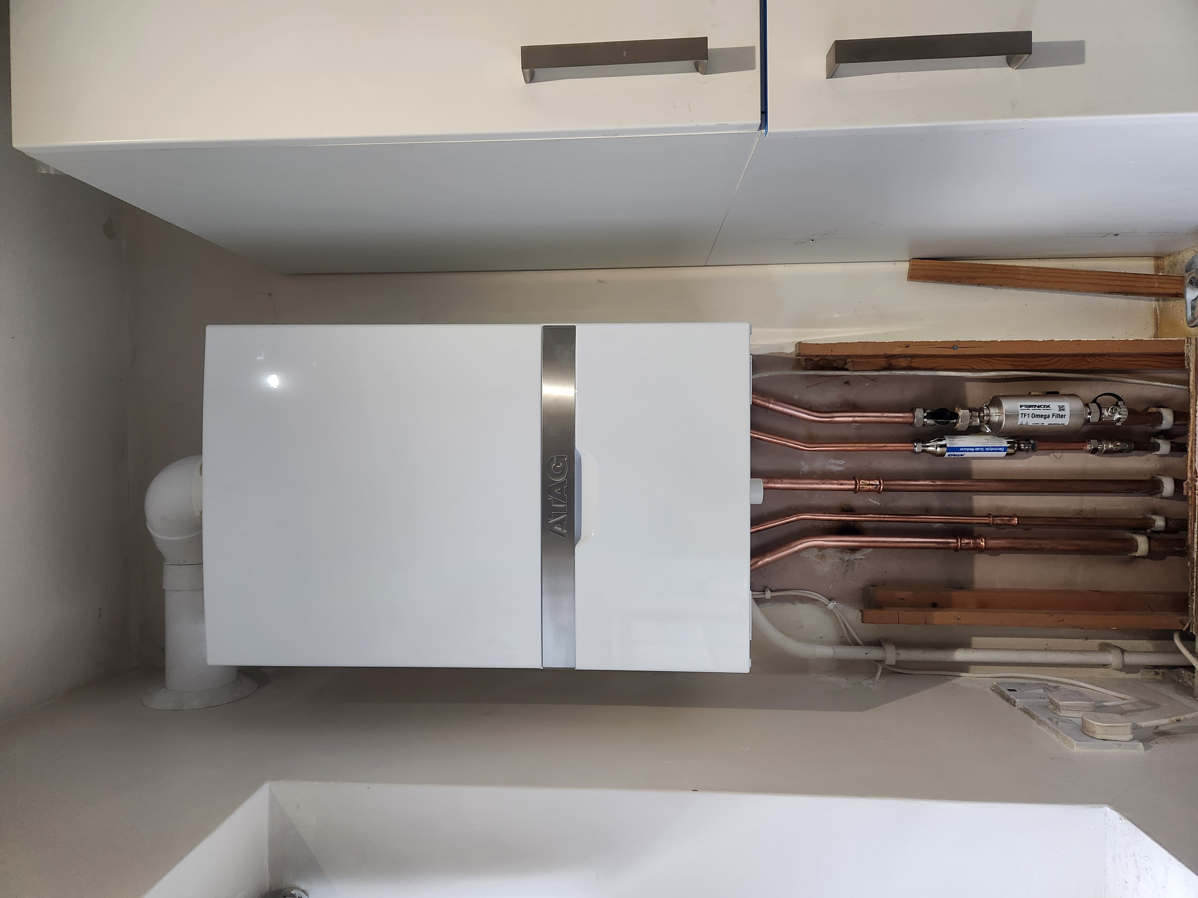 White ATAG combi boiler installed beneath kitchen cupboards with exposed copper pipework and magnetic filter.