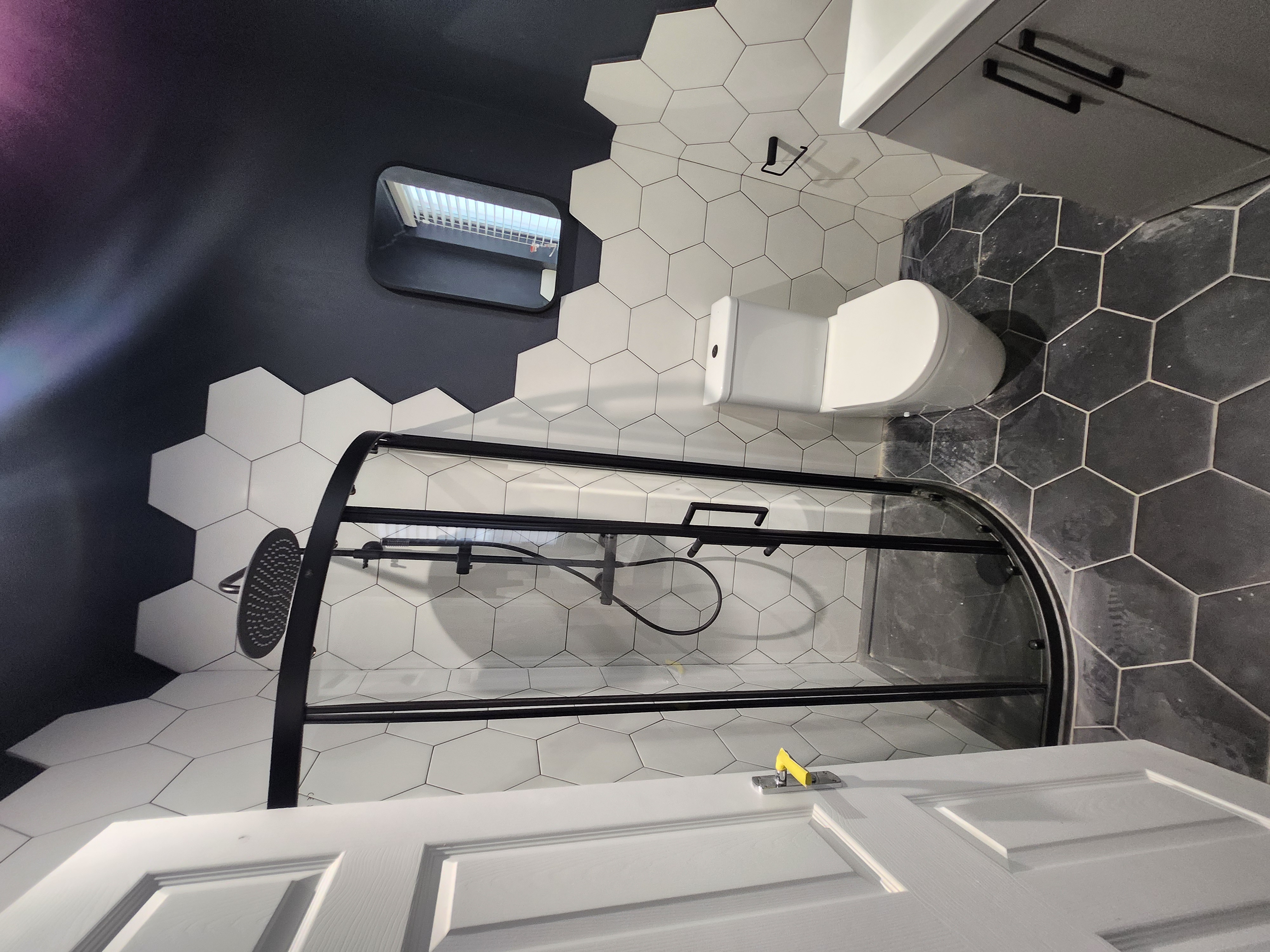 Modern bathroom interior with white hexagonal tiling, wall-mounted toilet, arched shower screen and black mixer tap.