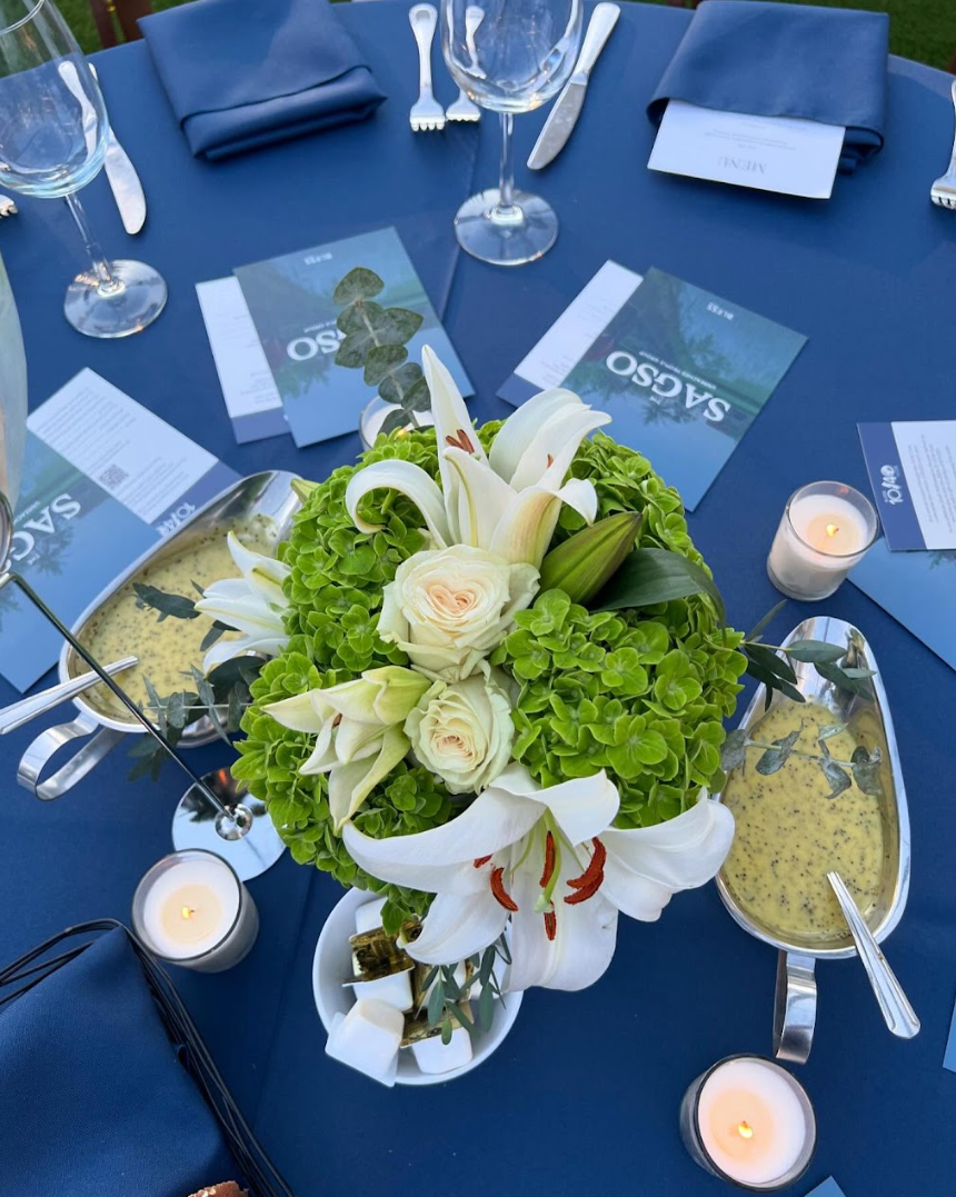 Elegant corporate dinner table with navy linens, white lily and green hydrangea centerpieces, printed menus, and glowing candles