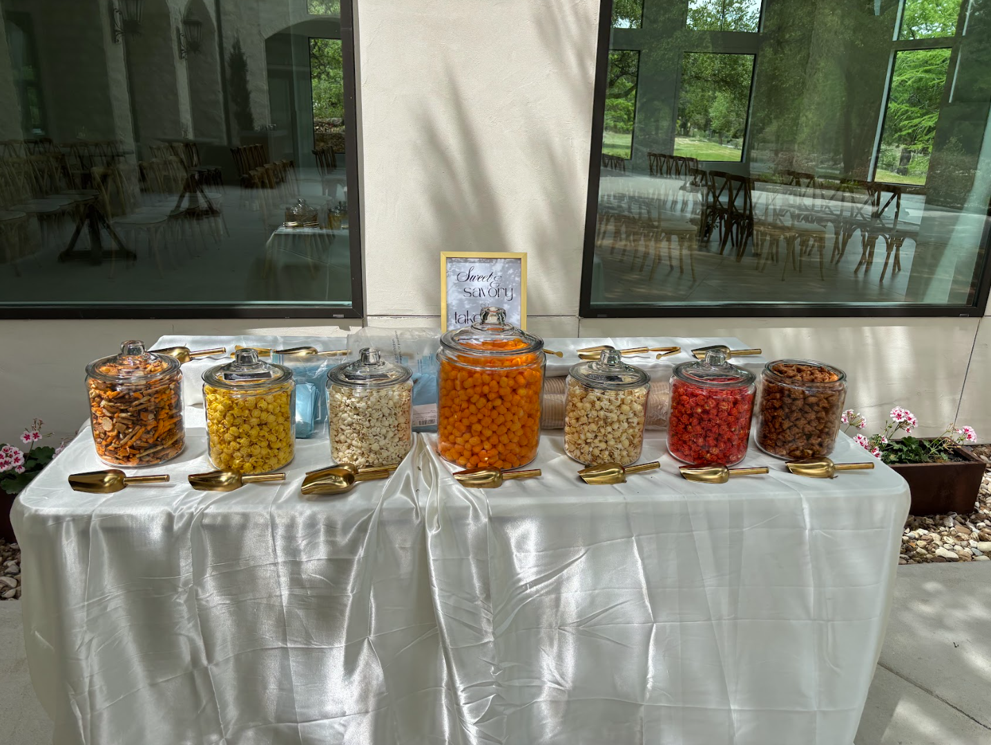 Elegant sweet and savory snack bar with glass jars of popcorn, pretzels, and treats on white linens with gold scoops
