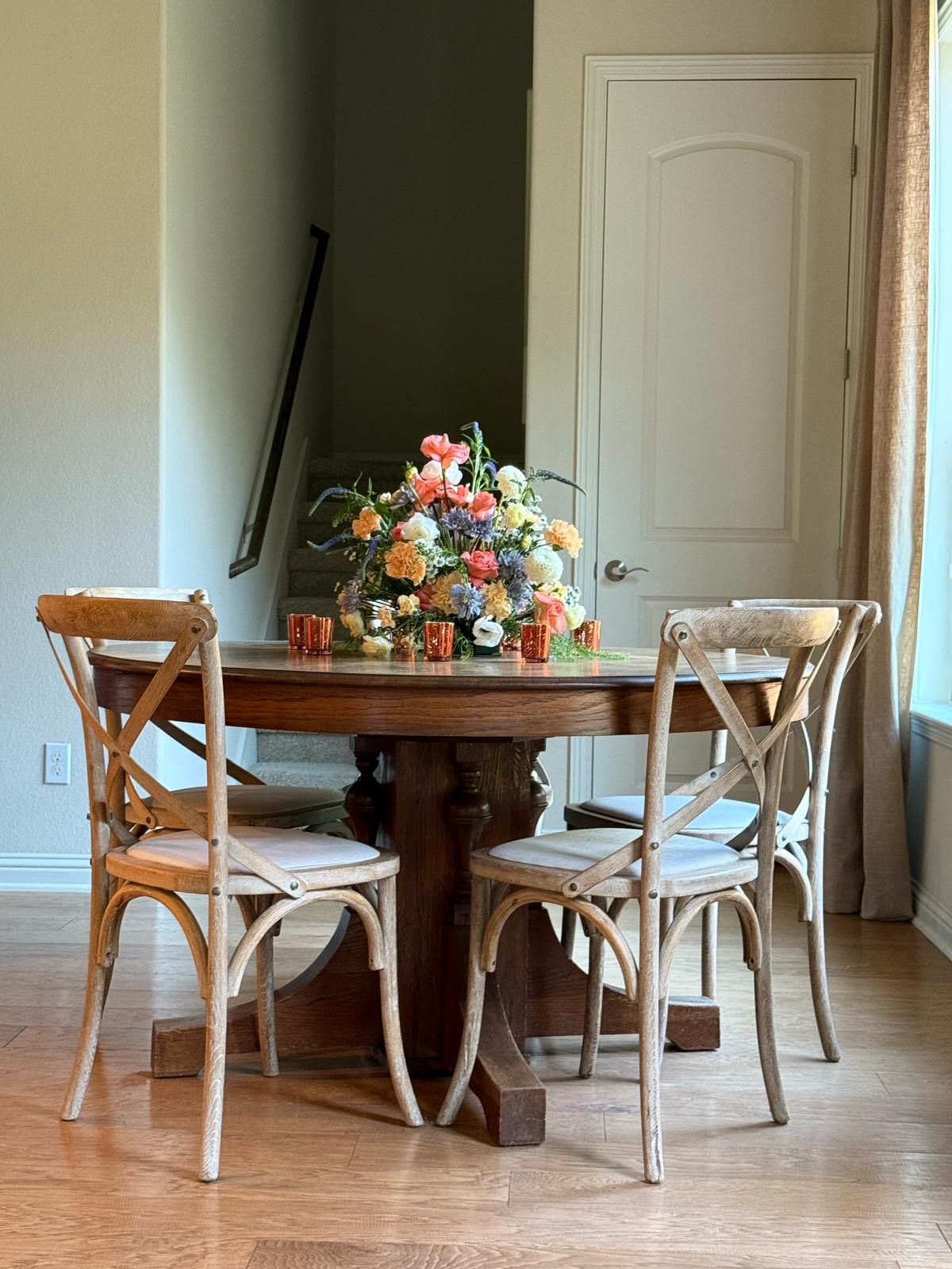 Round wooden pedestal table with rustic crossback chairs and vibrant floral centerpiece featuring peach, coral, and blue blooms