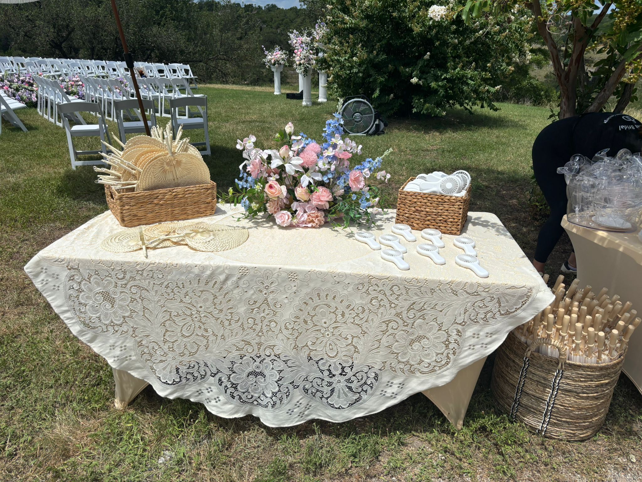 Beautiful outdoor wedding ceremony guest table with lace tablecloth, floral centerpiece, woven fans, and hand-held fans for guests