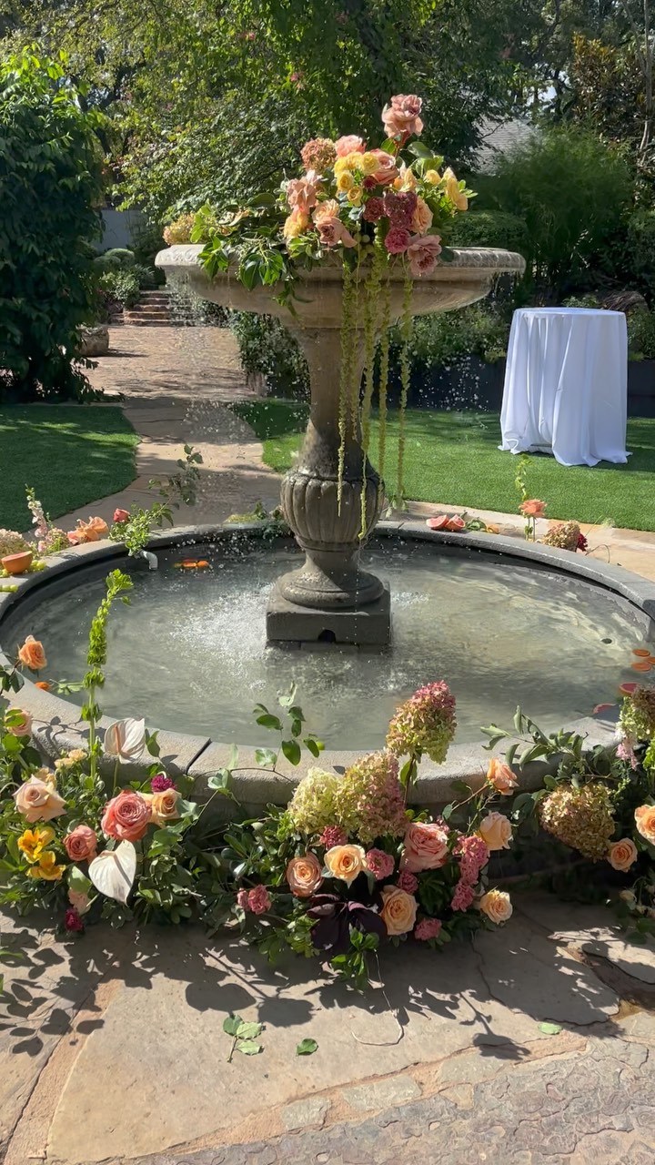 Stone fountain surrounded by lush peach and pink floral arrangements at outdoor wedding ceremony