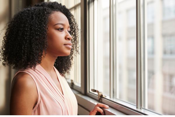 Woman standing by a window looking outside thoughtfully, representing reflection, uncertainty, and the feeling of putting life on hold while waiting for change.