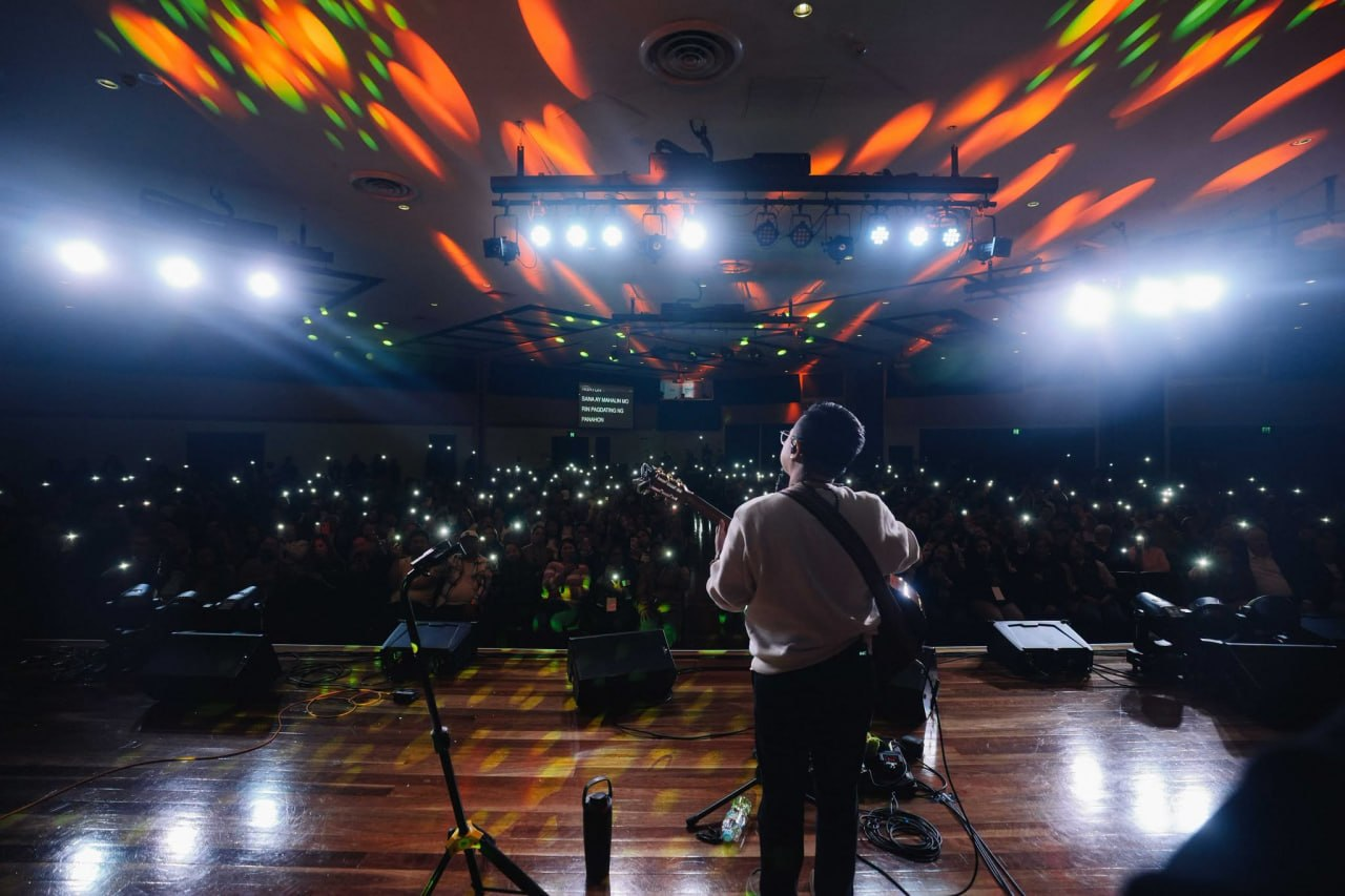 View from behind Ice Seguerra overlooking an auditorium lit by a sea of phone flashlights, surrounded by vibrant amber and green overhead lighting.