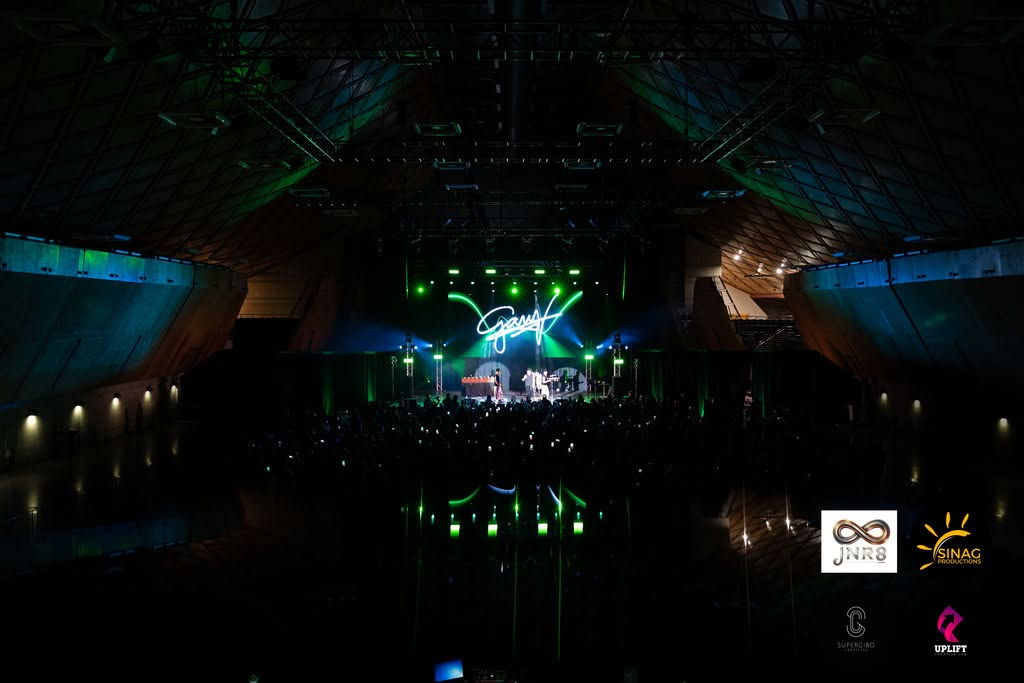 Wide arena view of the majestic concert venue lit in emerald green with Gary V performing on the grand stage for thousands of fans.