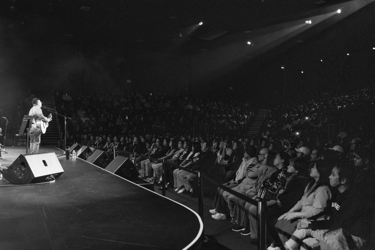 Side angle of Ice Seguerra playing guitar from stage left, looking out over a packed tiered theatre audience immersed in the live concert experience.