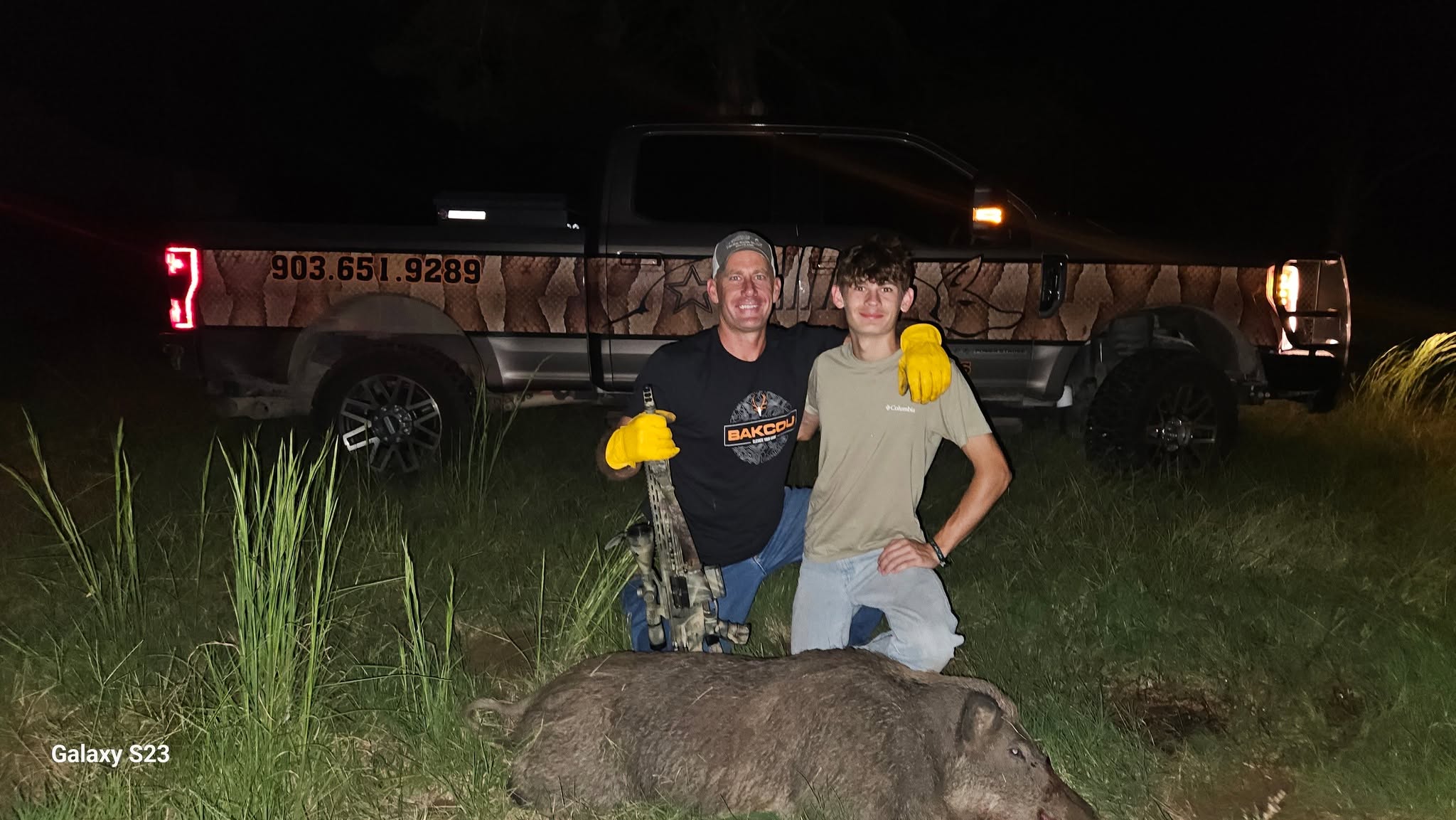 Father and son posing at night in front of Lone Star Wildlife Solutions branded truck with large captured feral boar on ground