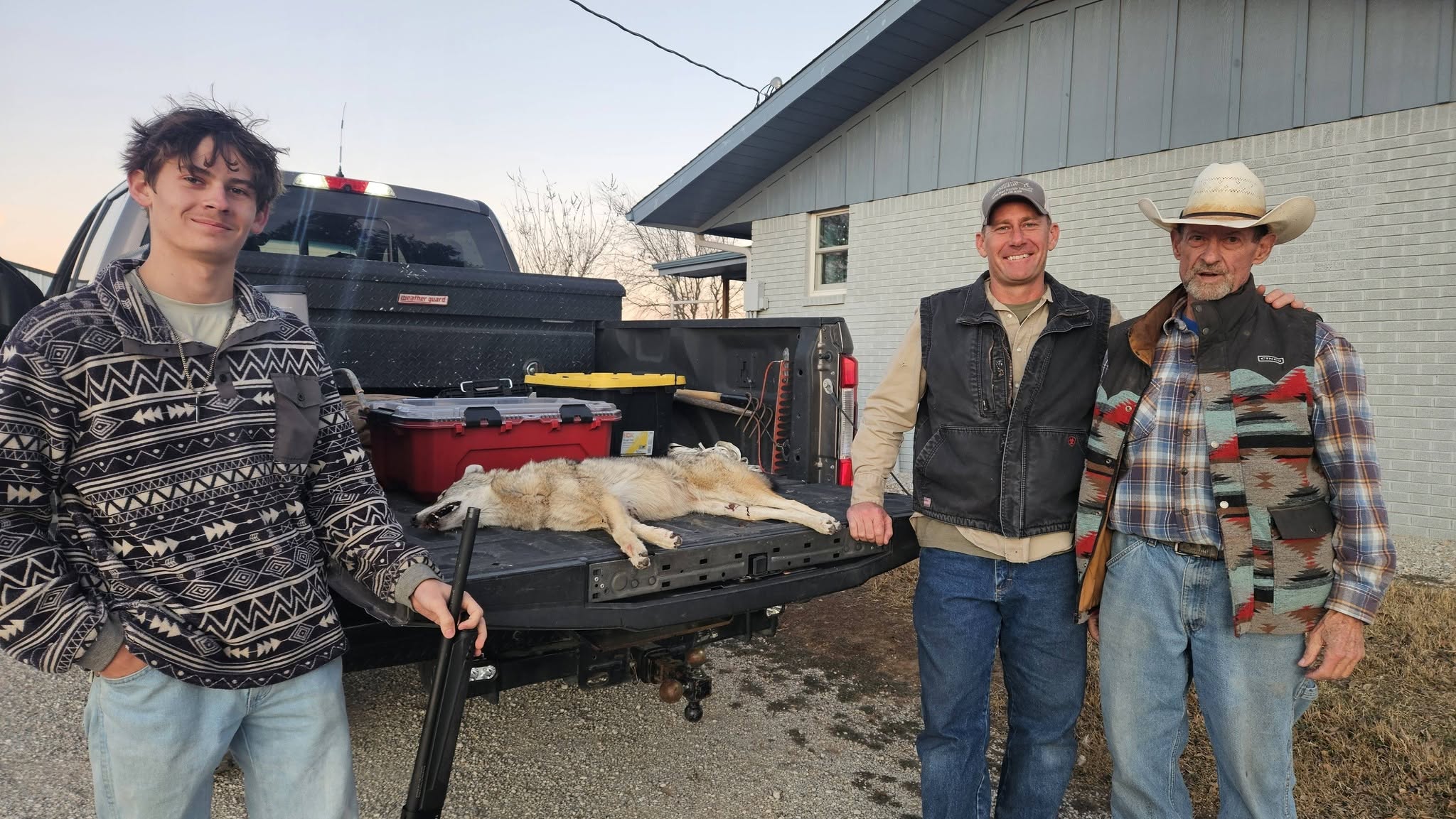 Three men standing next to black pickup truck tailgate with captured coyote laid out full length young man in patterned jacket man in vest and cap older man in cowboy hat and plaid vest house in background