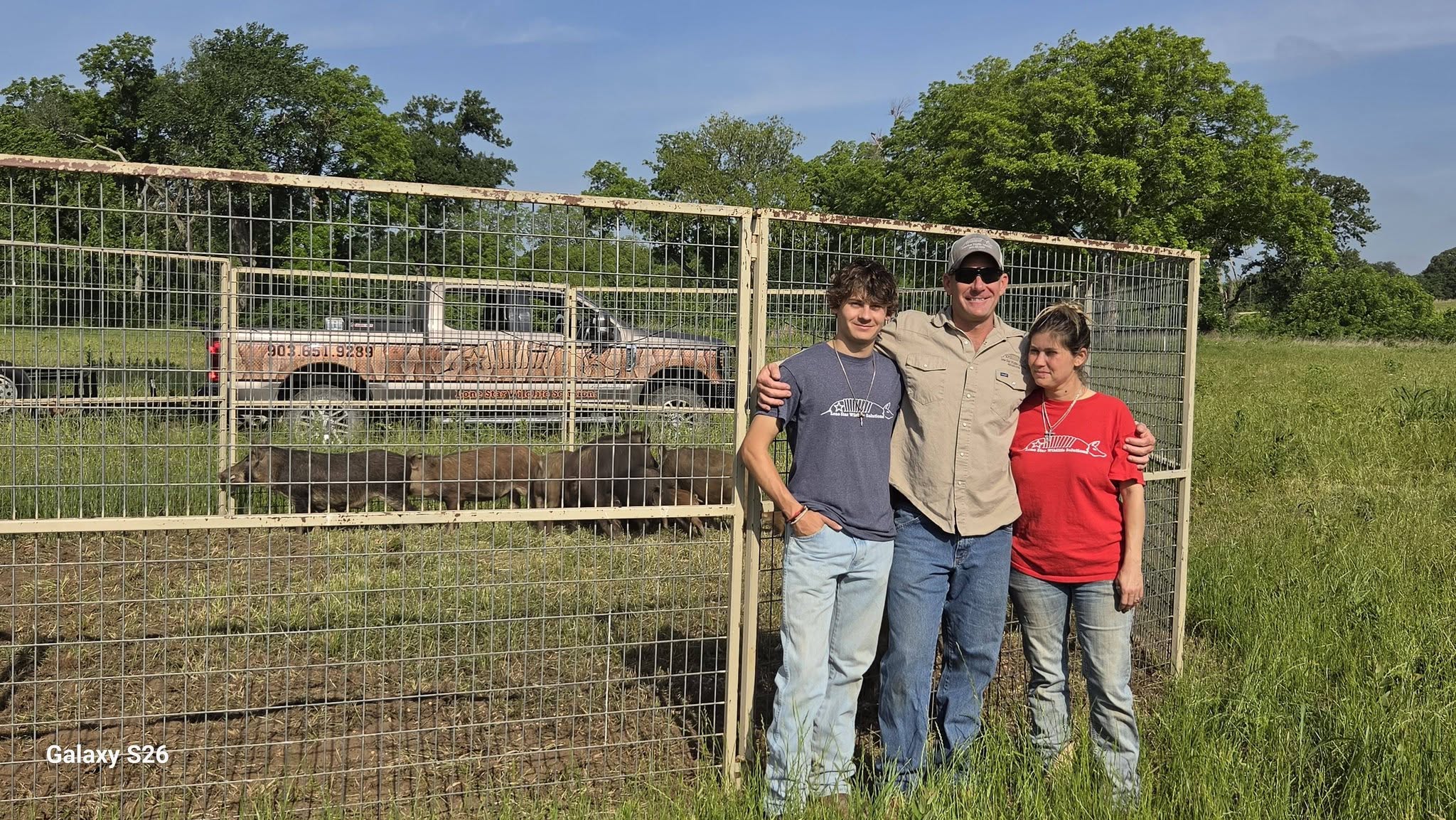Family of three — young man in blue t-shirt, man in tan work shirt and cap, woman in red t-shirt — standing together in front of wire corral fence with Lone Star Wildlife Solutions truck and feral hogs behind them on grassy field