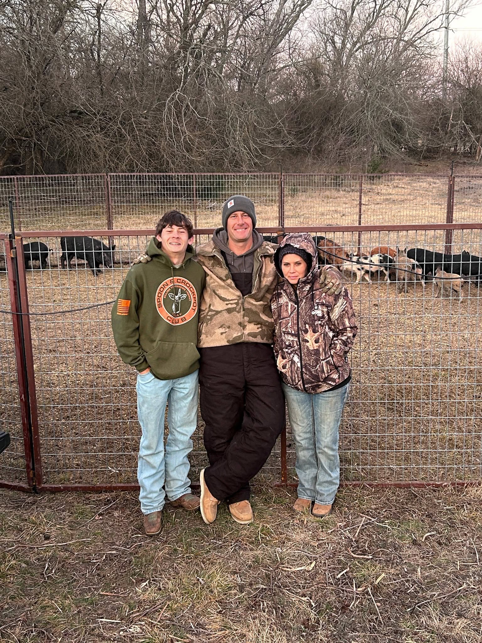 Family of three — father in camo jacket, son in green hoodie, mother in camo jacket — standing together in front of wire corral fence with feral hogs visible inside enclosure