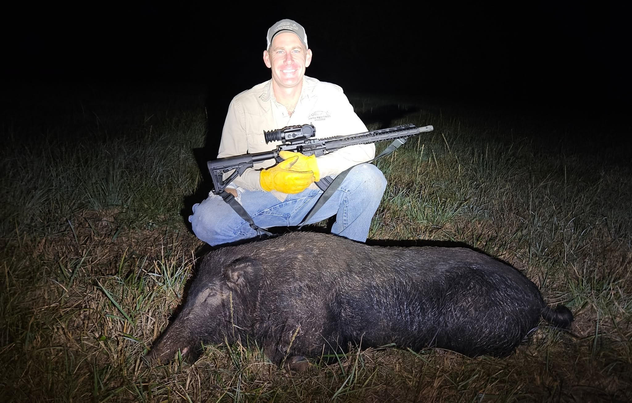 Lone Star Wildlife Solutions owner kneeling at night with night vision rifle next to large captured trophy feral boar on grass