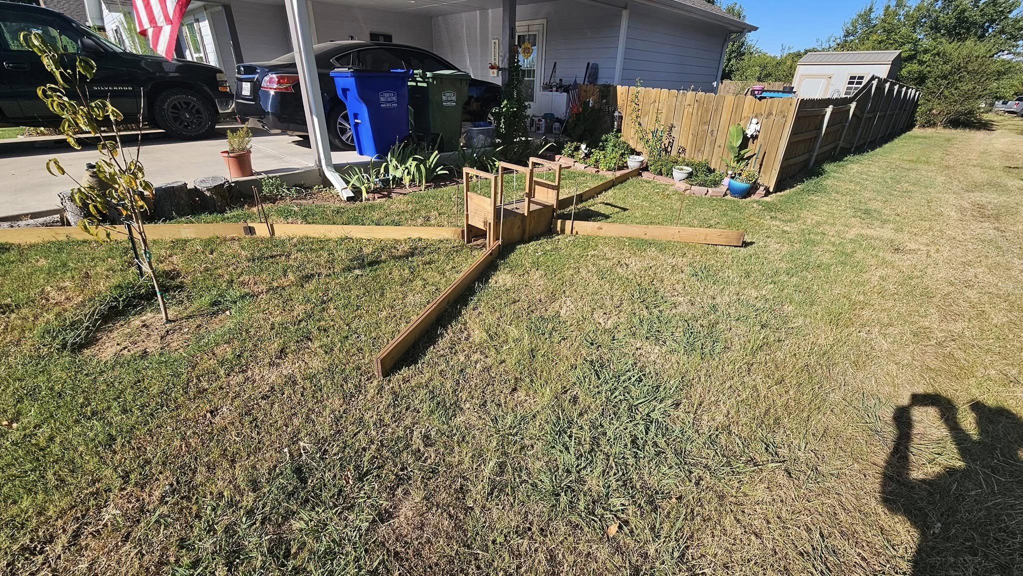 Residential backyard showing wooden armadillo trap enclosure set up on grass near house foundation with garden beds and fence visible