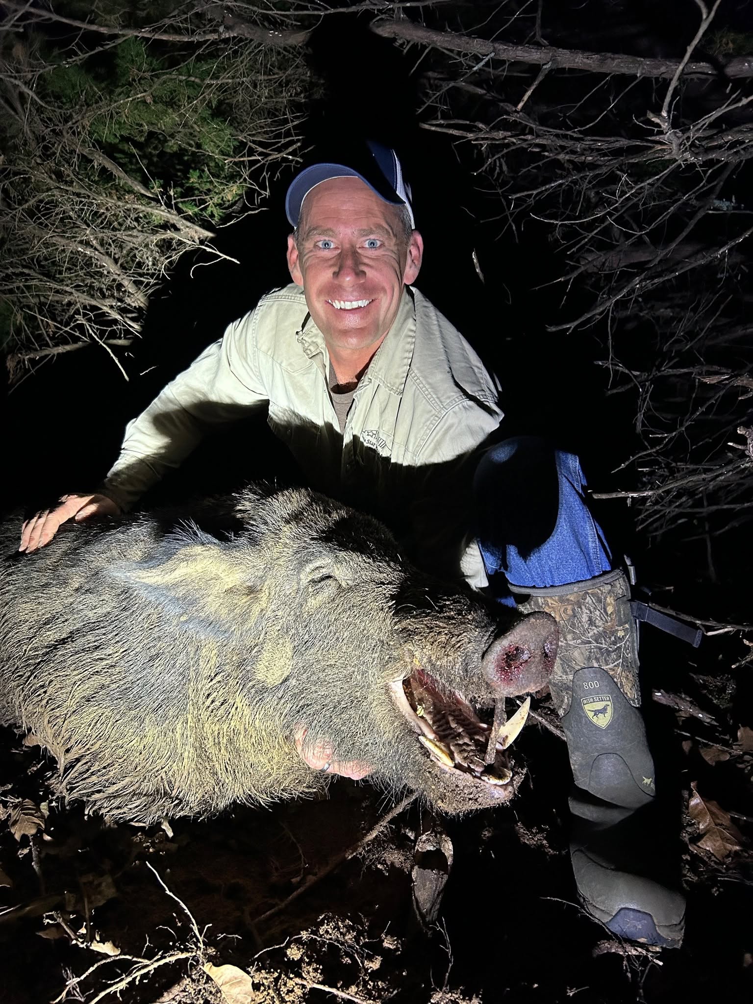 Lone Star Wildlife Solutions technician kneeling at night in brush with large captured trophy feral boar showing prominent tusks and open mouth