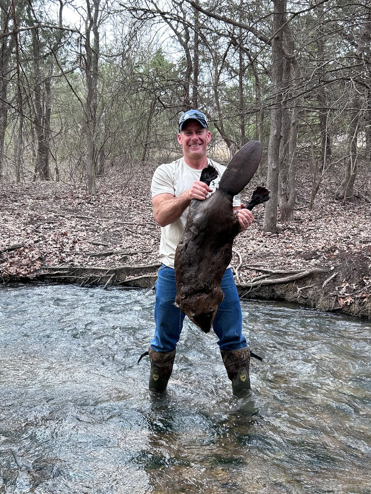 Man standing knee-deep in forest stream wearing rubber boots holding large captured beaver upright showing full body size flat tail and webbed hind feet