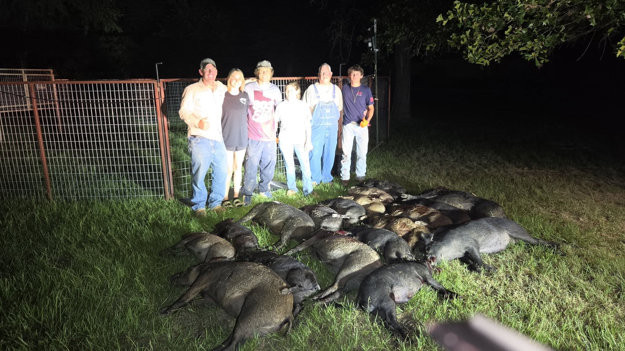 Large group of seven people including families standing behind wire fence at night with entire captured feral hog sounder of 20+ hogs laid out in rows on grassy field
