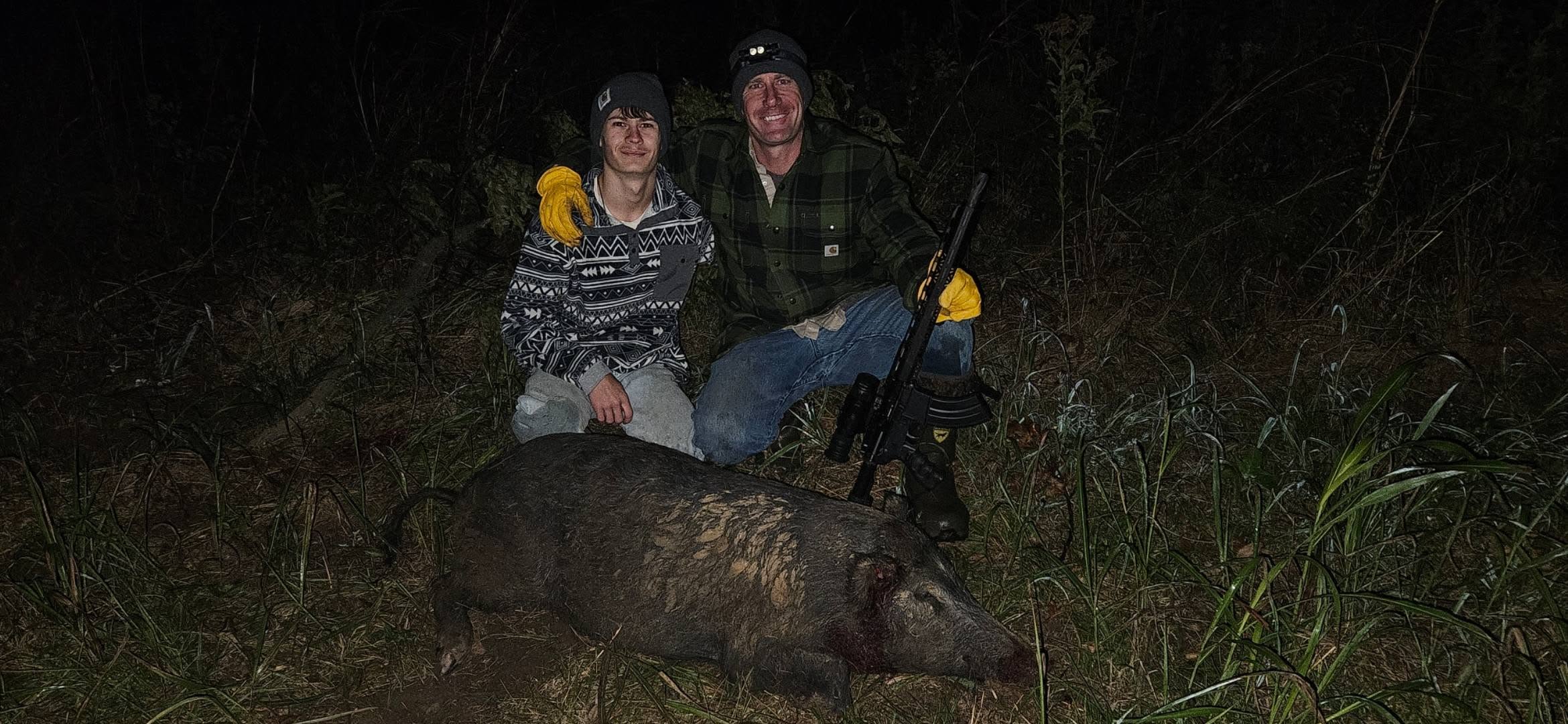 Two Lone Star Wildlife Solutions team members kneeling at night in tall grass with large captured feral boar on ground between them