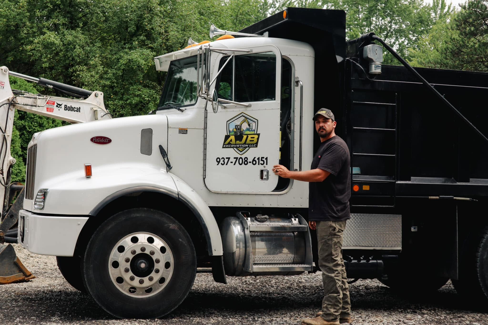 Andrew owner of AJB Excavation LLC standing with branded Peterbilt dump truck and Bobcat excavator fleet