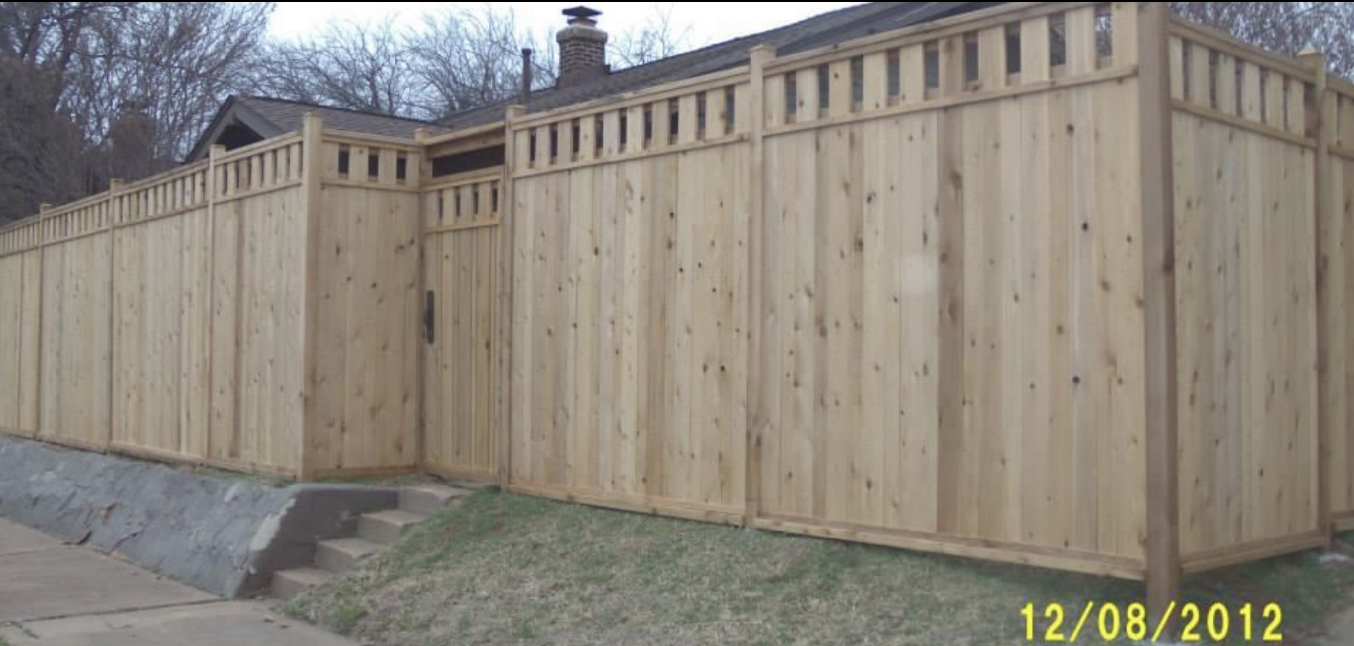 Custom Western Red Cedar wood privacy fence with vertical top spindle header and matching walk gate along stone retaining wall.
