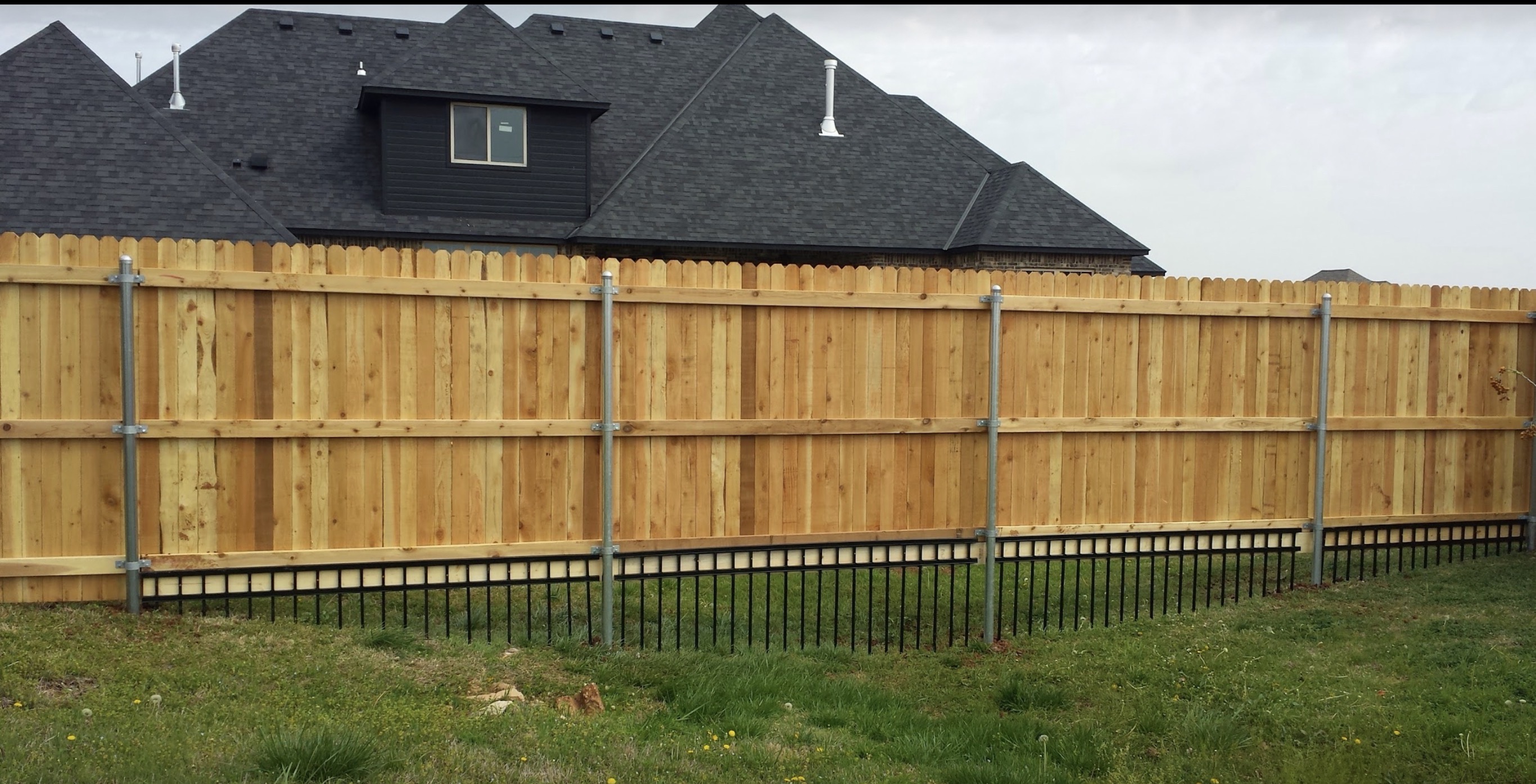 Western Red Cedar privacy fence constructed on heavy-gauge steel posts with an ornamental iron bottom barrier along grass lawn.