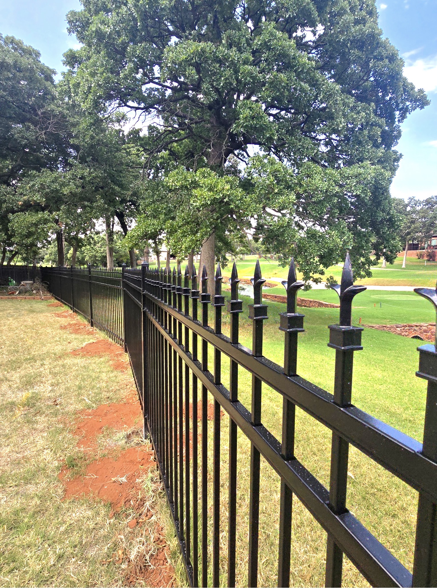 Close-up perspective of black ornamental steel fence pickets topped with decorative triad finials bordering a golf course.