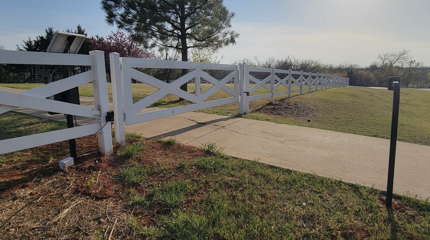 White crossbuck ranch style gate with solar powered automatic opener installed along paved walk.